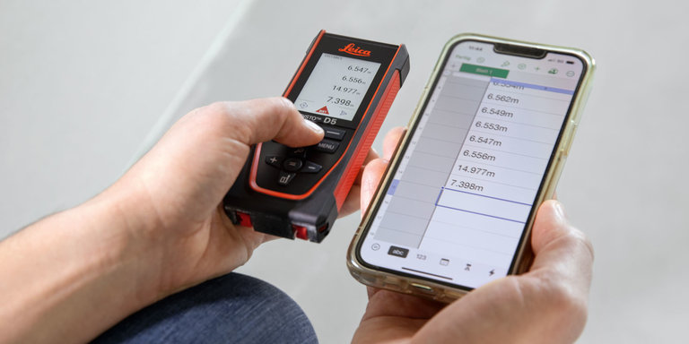 A person holds a Leica DISTO D5 in his left hand and a smartphone showing a table in his right hand and transfers measurement data from the laser measure to the smartphone