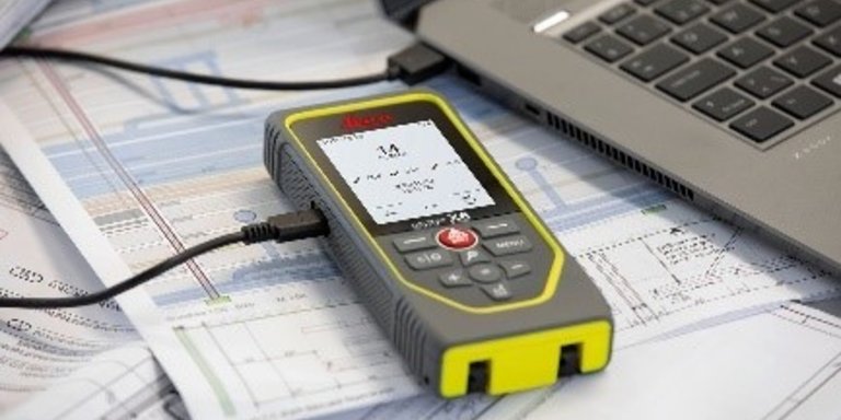 Leica DISTO X6 laser measure lies on drawings next to a laptop, transferring measurement reports to the laptop via a USB cable. 